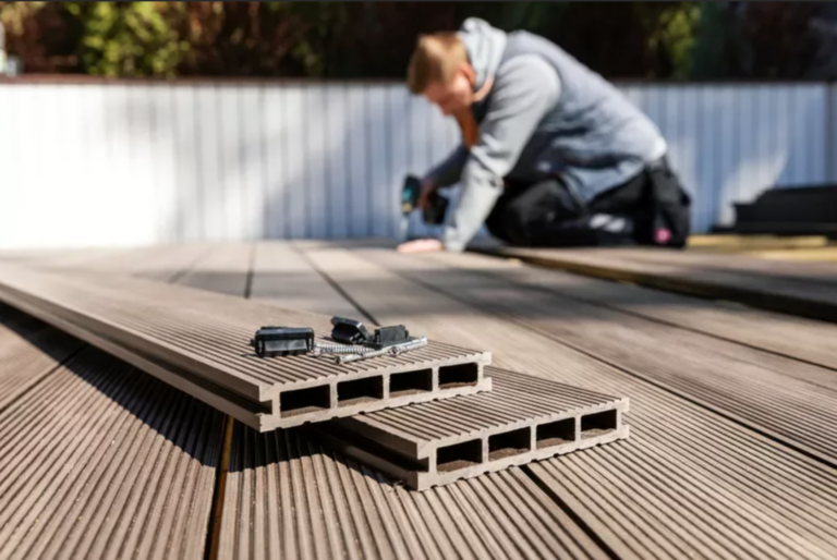 A man is setting up an outdoor wooden terrace with gray wood-plastic beige square planks on the ground. Next to him, there's a drill and some accessories lying around. The background shows white walls and a blue sky. The image was captured using a Canon EOS R5 camera with a wide-angle lens at an aperture value of f/20. Bright daylight illuminates the scene. --ar 67:44