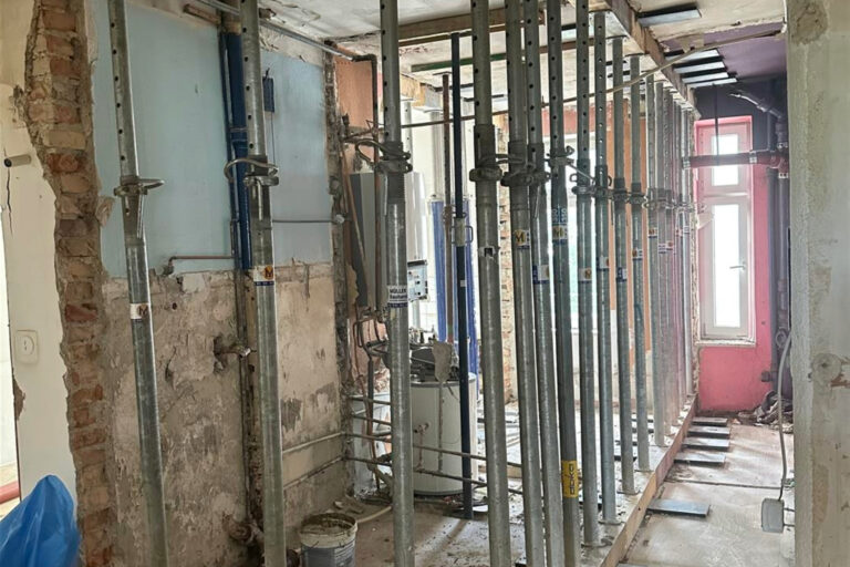 A photo of the interior wall being nestled with steel scaffolding and metal pipes, showing an industrial bathroom under construction in a London home. The room has partially plastered walls and a painted ceiling. There's one large window on the right side. This space will be transformed into a modern, open-plan kitchen-dining-living area, capturing the essence of transformation from old to new. --ar 107:72
