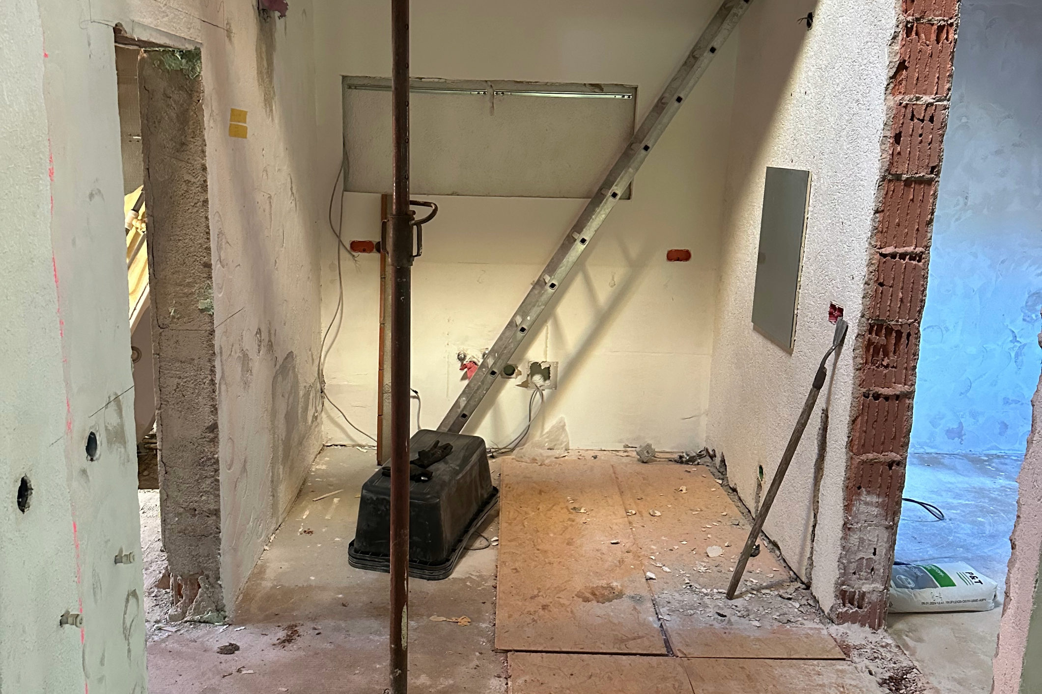 A photo of an empty room with walls and floor that have been sectioned off for electrical work, showing the open door leading to another part of the home. On one side, there is electrical equipment on the ground next to a ladder leaning against the wall. Some pipes and wires are visible in the background, and a window or small opening can be seen in the top left corner of the picture. --ar 31:21